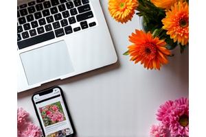 A smartphone with a social media feed displaying beautiful floral designs, surrounded by fresh flowers and a laptop, signifying digital marketing.